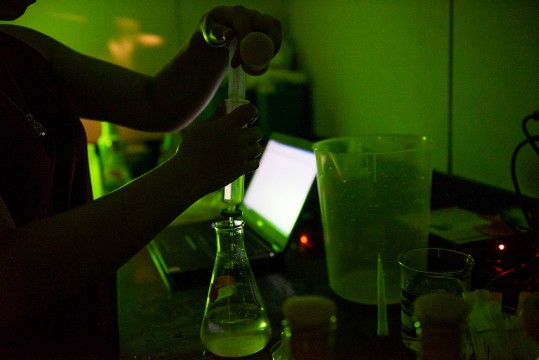 A scientist measures a liquid before pouring it in an erlenmeyer in a dimly lit laboratory bathed in green light.