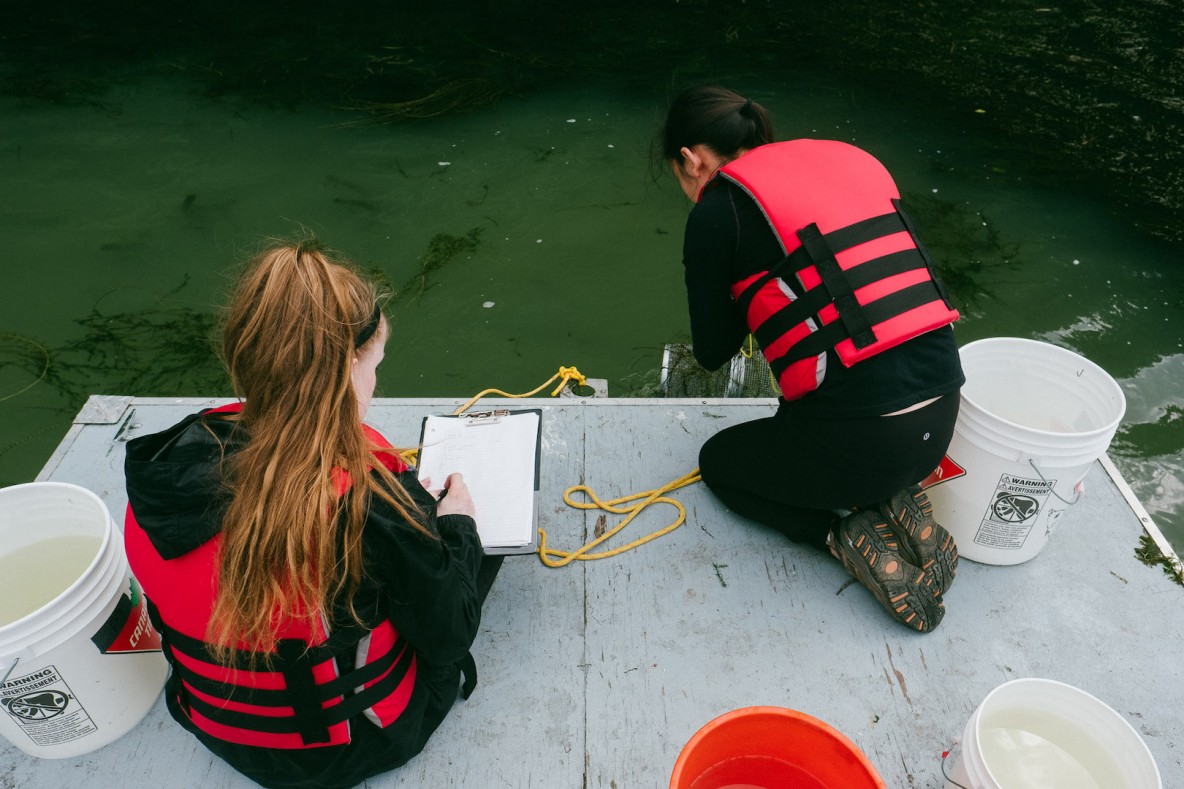 Two students are on a pontoon on Lac Hertel. One is taking notes while the other is pulling a fish trap out of the water.