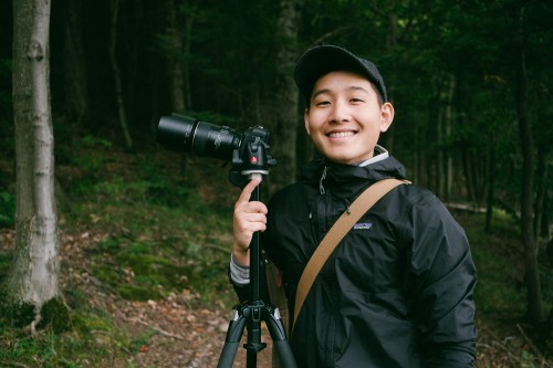 A smiling man is in the forest with a camera on a tripod