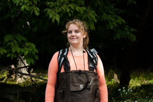 Portrait of a woman wearing waders on the shore of a lake.