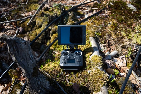 Une télécommande de drône arborant plusieurs boutons et un écran est placée sur le sol de la forêt