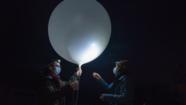 Two scientists are standing in the dark with frontal lamps on their foreheads. Raindrops are illuminated by the lamps. On the left, the researcher hold a large white balloon with one hand and a rectangular device with the other. The two devices are joined by a rope.