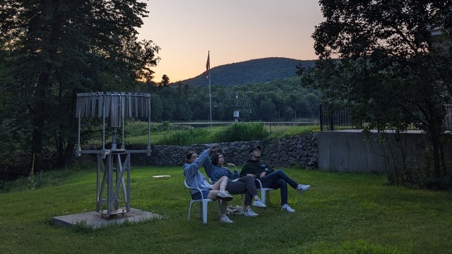 Trois stagiaires assis sur des chaises de jardin en plastique, observant quelque chose hors champ. L’une d’eux pointe du doigt ce qu’ils regardent. En arrière-plan, le coucher de soleil est visible au-dessus de la montagne.