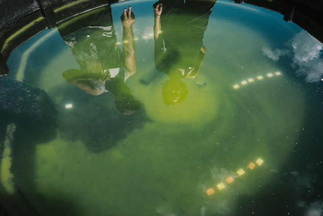 Two researchers are reflected on the surface of the water in a black artificial pond