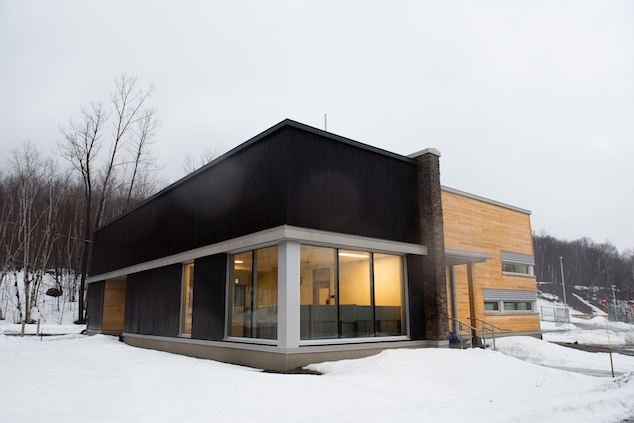 A modern looking building made of wood with large windows can be seen against a winter forested backdrop.