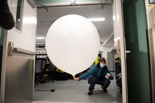 A scientist wearing a blue coat, a hat, and a frontal lamp is crouching to help a very large white balloon fit through a doorway.