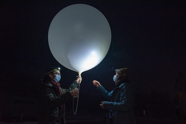 Two scientists are standing in the dark with frontal lamps on their foreheads. Raindrops are illuminated by the lamps. On the left, the researcher hold a large white balloon with one hand and a rectangular device with the other. The two devices are joined by a rope.