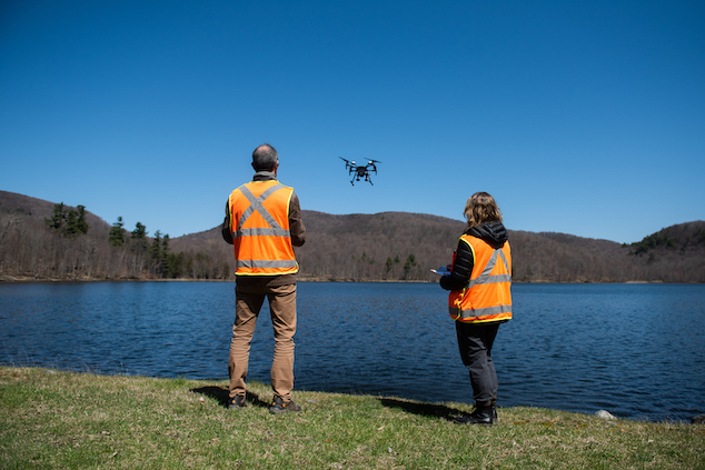 Photo of two people facing away from the camera towards a drone flying over a lake