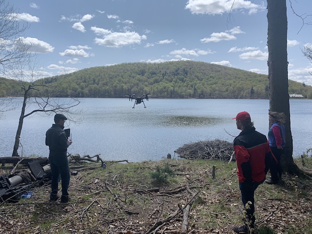 Des membres du personnel observent un drone planant au-dessus du lac Hertel à partir de la rive. 