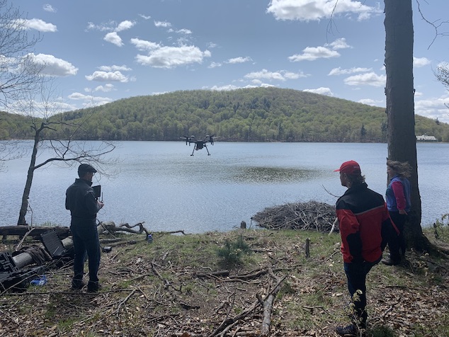 A drone hovers low over Lac Hertel. The pilot and two employees monitor it from the shore. 